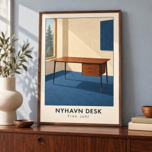 May include: A framed art print featuring a wooden desk with three drawers and dark legs set against a blue floor and cream walls. The text NYHAVN DESK and Finn Juhl is printed at the bottom. A window shows a view of pine trees outside.