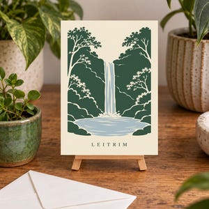 K&ouml;nnte beinhalten: Ein rechteckiger Kunstdruck auf einer kleinen Holzstaffelei mit einer stilisierten Illustration eines Wasserfalls, der in ein Becken st&uuml;rzt, umgeben von dunkelgr&uuml;nen B&auml;umen auf cremefarbenem Grund, mit dem Text LEITRIM unten in der Mitte.