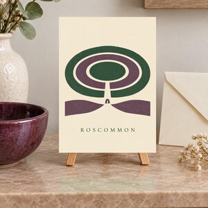 May include: A rectangular print on a small wooden easel featuring a cream background with a dark green and purple abstract circular design. Below the graphic, the word ROSCOMMON is printed in dark green capital letters in a simple font.