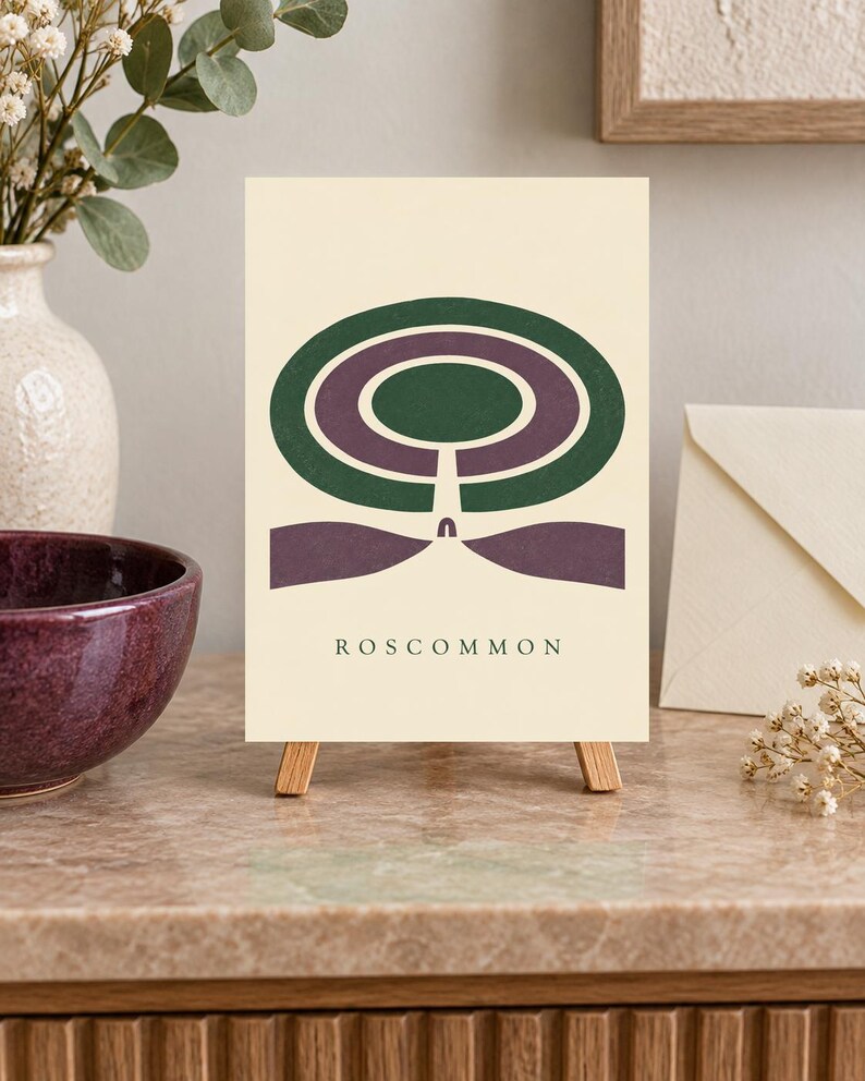 May include: A rectangular print on a small wooden easel featuring a cream background with a dark green and purple abstract circular design. Below the graphic, the word ROSCOMMON is printed in dark green capital letters in a simple font.