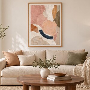 May include: A framed abstract wall art print featuring organic shapes in muted tones of dusty pink, terracotta, cream, olive green, and a dark navy blue arc, displayed in a light wood frame with a white mat border on a neutral-colored wall.