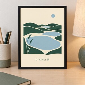May include: A framed wall art print featuring a stylized landscape illustration of rolling dark green hills and light blue lakes under a pale yellow sky with a small blue sun. The word CAVAN is printed in dark green capital letters at the bottom.