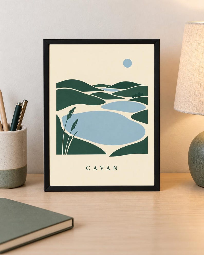 May include: A framed wall art print featuring a stylized landscape illustration of rolling dark green hills and light blue lakes under a pale yellow sky with a small blue sun. The word CAVAN is printed in dark green capital letters at the bottom.