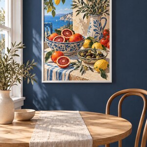 Op de afbeelding: Een ingelijste kunstdruk met een mediterrane sc&egrave;ne met een witte tafel met blauw-wit gedessineerde kommen gevuld met bloedsinaasappels, citroenen en olijven. Een blauw-witte kan met olijftakken staat in de buurt, uitkijkend over een kustzee.