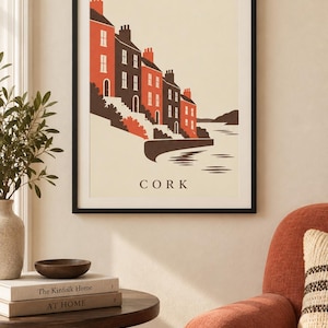 Op de afbeelding: Een ingelijste kunstdruk met een gestileerde illustratie van een rij huizen langs een waterkant in Cork. De gebouwen zijn weergegeven in tinten oranje en donkergrijs tegen een cr&egrave;mekleurige achtergrond, met het woord CORK eronder.