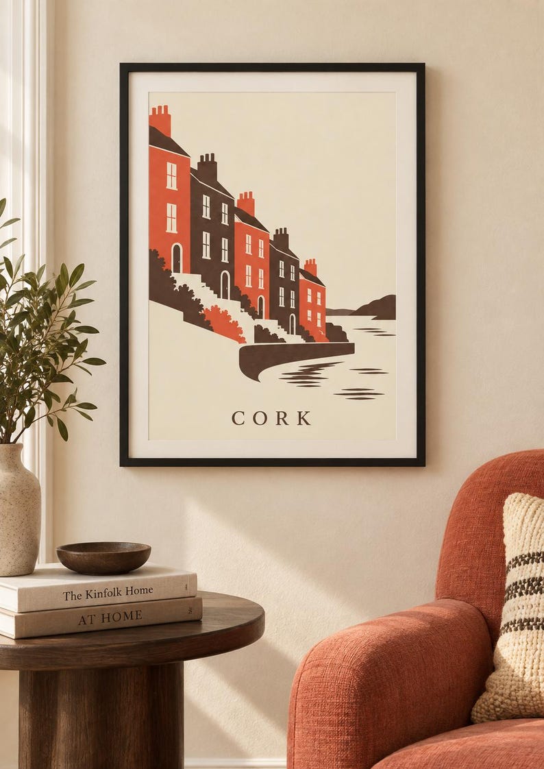 Op de afbeelding: Een ingelijste kunstdruk met een gestileerde illustratie van een rij huizen langs een waterkant in Cork. De gebouwen zijn weergegeven in tinten oranje en donkergrijs tegen een cr&egrave;mekleurige achtergrond, met het woord CORK eronder.