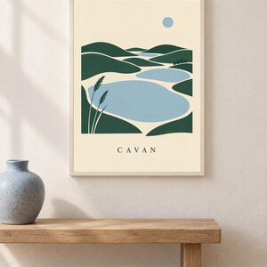 May include: A framed wall art print featuring a stylized landscape with dark green rolling hills, light blue bodies of water, and a small blue sun against a cream background. Two tall grass stalks are on the left. The text CAVAN is printed below.