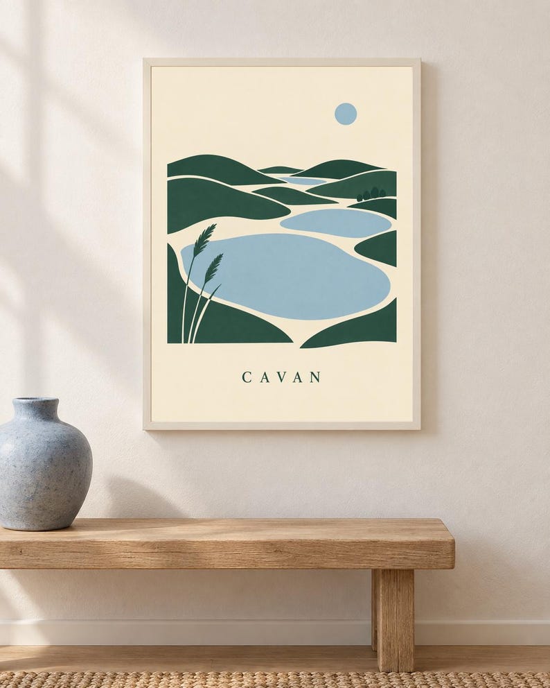 May include: A framed wall art print featuring a stylized landscape with dark green rolling hills, light blue bodies of water, and a small blue sun against a cream background. Two tall grass stalks are on the left. The text CAVAN is printed below.