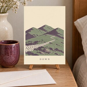 Pu&ograve; includere: Una stampa artistica rettangolare su un piccolo cavalletto in legno raffigurante un paesaggio con colline verdi, un muretto in pietra e ombre viola sotto un cielo color crema. La parola DOWN &egrave; stampata in lettere maiuscole nere in basso.