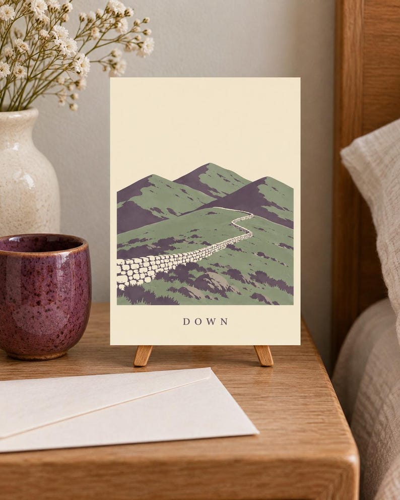 Pu&ograve; includere: Una stampa artistica rettangolare su un piccolo cavalletto in legno raffigurante un paesaggio con colline verdi, un muretto in pietra e ombre viola sotto un cielo color crema. La parola DOWN &egrave; stampata in lettere maiuscole nere in basso.