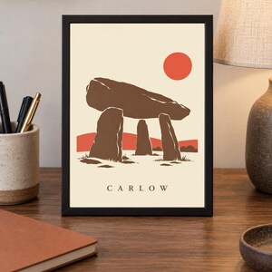 May include: A framed art print featuring a stylized illustration of the Brownshill Dolmen in Carlow, Ireland. The design displays brown megalithic stones against a cream background with a solid red sun and red hills. The text CARLOW is printed below.