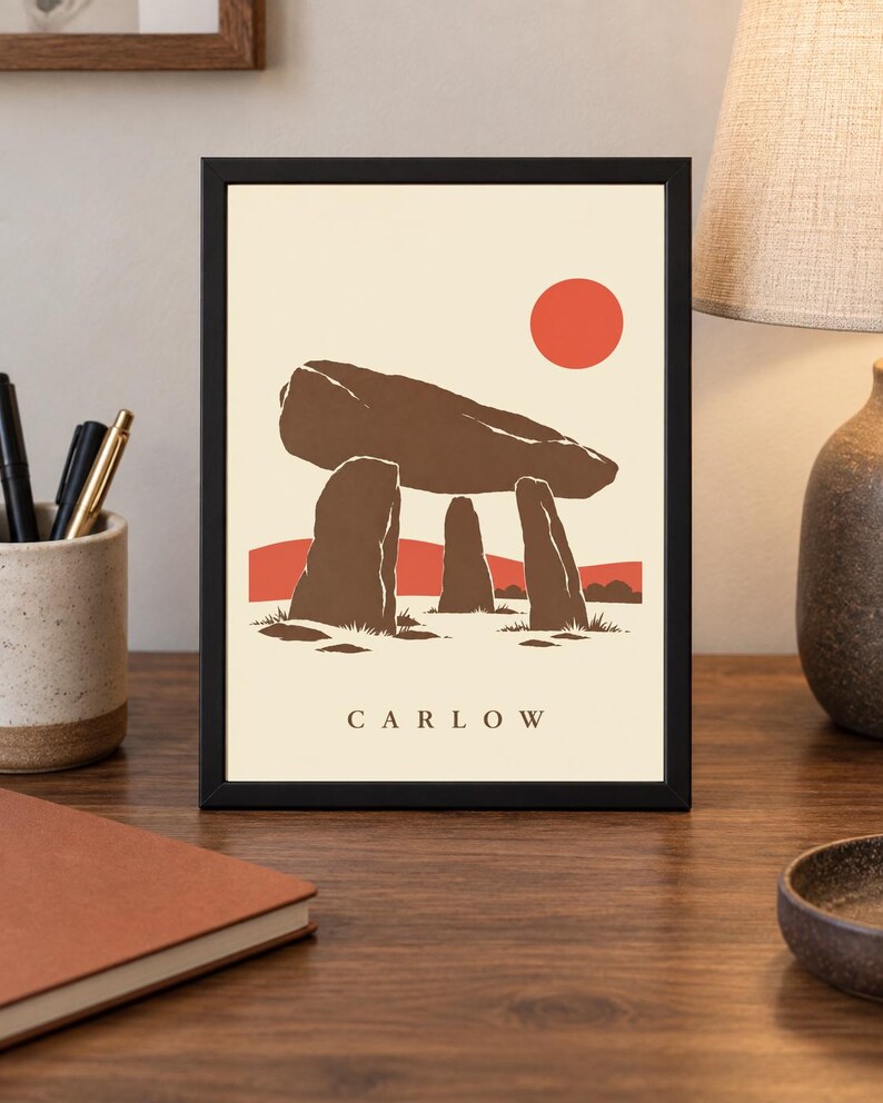 May include: A framed art print featuring a stylized illustration of the Brownshill Dolmen in Carlow, Ireland. The design displays brown megalithic stones against a cream background with a solid red sun and red hills. The text CARLOW is printed below.