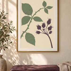 May include: A framed wall art print featuring a botanical illustration on a cream background. The artwork displays a green thorny stem with serrated leaves on the left and a cluster of dark purple rose hips on the right, all enclosed in a gold frame.