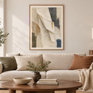 May include: A framed abstract wall art print featuring layered paper-like shapes in shades of cream, beige, and charcoal gray, with a single vertical gold accent mark. The composition includes soft, flowing lines and deep dark blue color blocks.