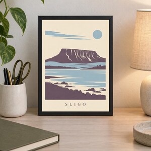 May include: A framed art print featuring a stylized landscape illustration of Sligo with a prominent flat-topped mountain, a body of water, and a pale blue sun against a cream background. The word SLIGO is printed in black at the bottom center.