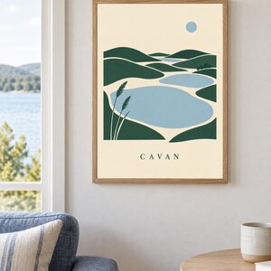 May include: A framed wall art print featuring a stylized landscape illustration of green rolling hills, light blue bodies of water, and a small blue circle representing the sun. The word CAVAN is printed in black capital letters at the bottom.