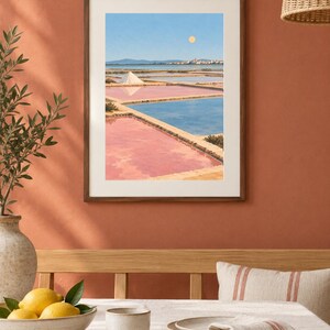May include: A framed wall art print depicting a landscape of salt pans with pink and blue water, a white salt mound, and a distant town under a pale sun in a clear blue sky. The artwork is enclosed in a simple dark wood frame with a white mat border.