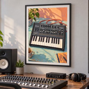 Op de afbeelding: Een ingelijste muurkunstprint met een zwarte Arturia MiniBrute analoge synthesizer met witte toetsen, zwarte knoppen en schuifregelaars, rustend op een blauw oppervlak met een rode kabel, een plant, een mok en een notitieboek in de compositie.