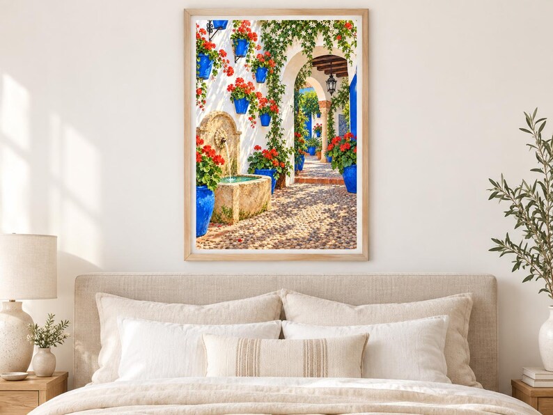 May include: A framed wall art print depicting a sunlit Mediterranean courtyard with a stone fountain, vibrant red geraniums in bright blue pots mounted on white walls, climbing green ivy, and a cobblestone path leading through an arched doorway.