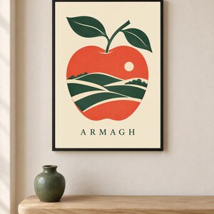 May include: A framed wall art print featuring a stylized red apple graphic on a cream background. Inside the apple shape, there is a landscape illustration of green rolling hills and a small white sun. The text ARMAGH is printed in dark green.