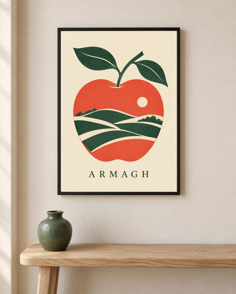 May include: A framed wall art print featuring a stylized red apple graphic on a cream background. Inside the apple shape, there is a landscape illustration of green rolling hills and a small white sun. The text ARMAGH is printed in dark green.