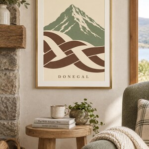 K&ouml;nnte beinhalten: Ein gerahmter Kunstdruck mit einem stilisierten gr&uuml;nen Berggipfel mit wei&szlig;en Schneeakzenten &uuml;ber einem abstrakten braun-cremefarbenen keltischen Knotenmuster. Das Wort DONEGAL ist in schwarzen Gro&szlig;buchstaben auf dem cremefarbenen Hintergrund gedruckt.