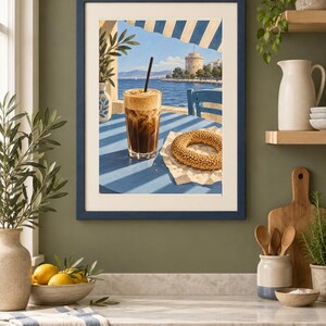 May include: A framed art print featuring a painting of a glass of iced coffee with a black straw and a sesame seed bagel on a blue table. The scene depicts a coastal view with a white tower in the distance under a blue sky and striped awning.