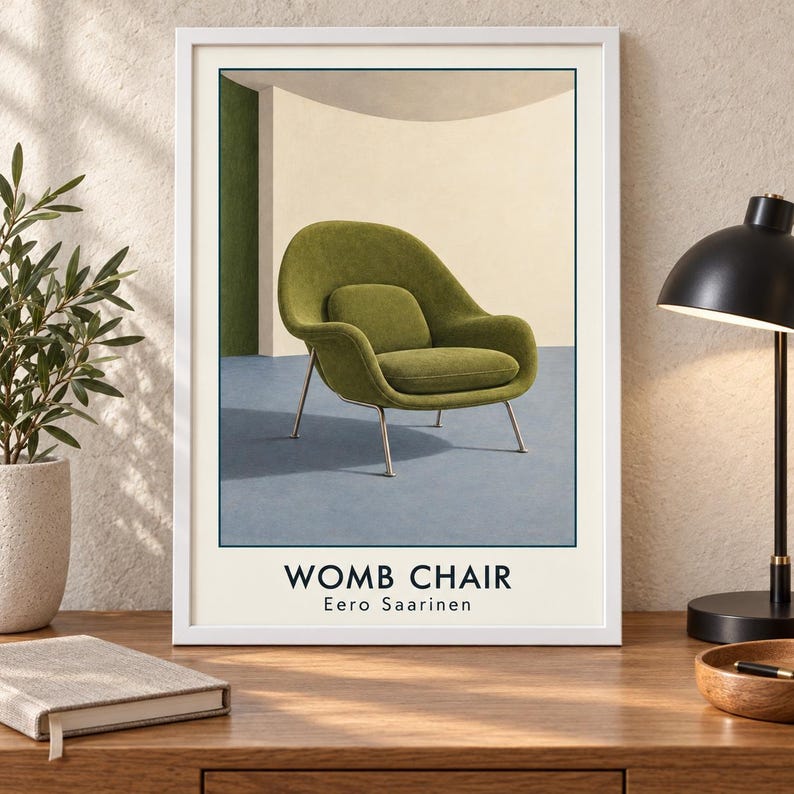 May include: A framed wall art print featuring a green Womb Chair designed by Eero Saarinen. The illustration shows the chair with thin metal legs against a cream and green background. The text WOMB CHAIR and Eero Saarinen appears below.