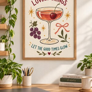 May include: A framed wall art print featuring a cocktail glass with a cherry, a cluster of purple grapes, and a pink ribbon. The text reads A TOAST TO THE LOVELY THINGS and LET THE GOOD TIMES GLOW, accented by yellow starbursts and a small red heart.