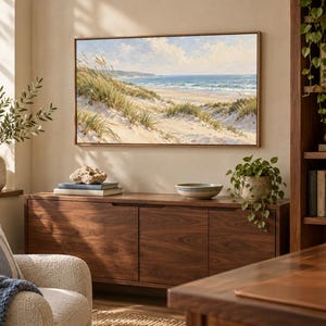 May include: A rectangular landscape painting in a thin wooden frame depicting a coastal scene with sand dunes, tall beach grass, and a blue ocean under a cloudy sky, mounted on a beige wall above a dark wood sideboard cabinet.
