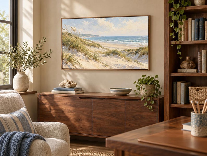 May include: A rectangular landscape painting in a thin wooden frame depicting a coastal scene with sand dunes, tall beach grass, and a blue ocean under a cloudy sky, mounted on a beige wall above a dark wood sideboard cabinet.