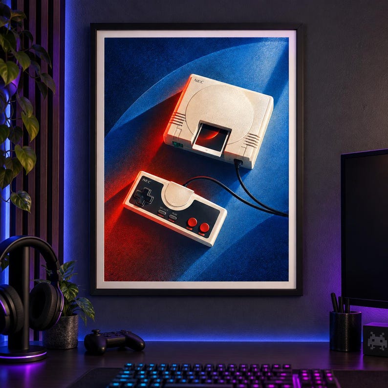 May include: A framed wall art print featuring a vintage NEC video game console and a matching wired controller. The console is off-white with a disc drive slot, and the controller has a black directional pad, two red buttons, and select and run labels.