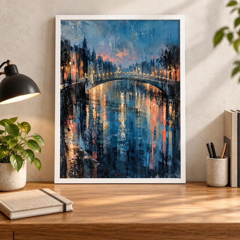 Op de afbeelding: Een ingelijste muurkunstprint met een stadsgracht bij schemering. Het schilderij toont een stenen brug, gebouwen langs het water en oranje straatlantaarns die reflecteren in het diepblauwe water onder een zachte roze en blauwe lucht.