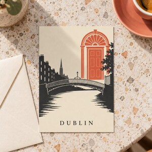 K&ouml;nnte beinhalten: Eine rechteckige Postkarte mit einer grafischen Illustration der Skyline von Dublin, einer Br&uuml;cke &uuml;ber einen Fluss und einer markanten orangefarbenen T&uuml;r im georgianischen Stil. Das Wort DUBLIN ist in schwarzen Gro&szlig;buchstaben unten aufgedruckt.