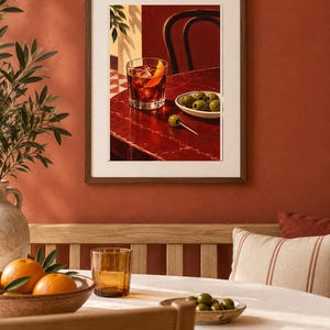 May include: A framed wall art print depicting a dark red table surface with a glass of amber liquid containing ice and an orange peel, a small white bowl filled with green olives, and a single green olive on a toothpick, set against a dark background.