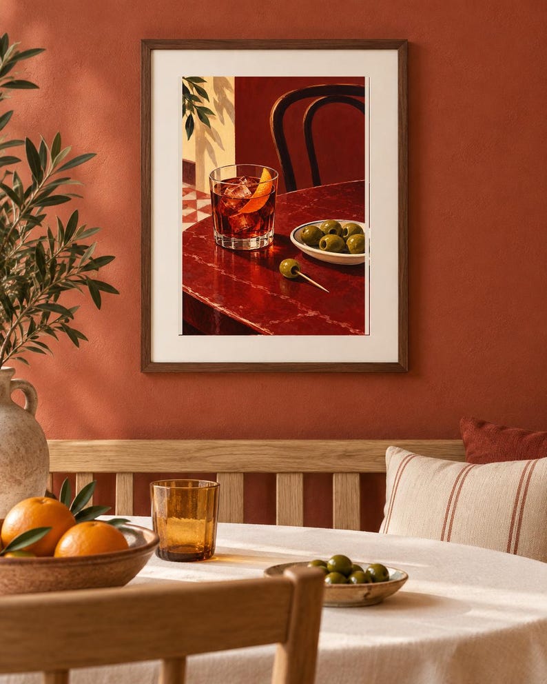 May include: A framed wall art print depicting a dark red table surface with a glass of amber liquid containing ice and an orange peel, a small white bowl filled with green olives, and a single green olive on a toothpick, set against a dark background.