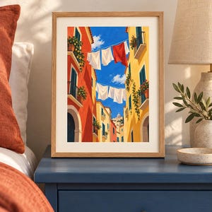 Op de afbeelding: Een ingelijste kunstdruk toont een smalle straat tussen hoge oranje en gele gebouwen onder een helderblauwe lucht. Tussen de gebouwen hangt wasgoed aan lijnen, waaronder witte en rode stoffen. Potplanten sieren de balkons van de gebouwen.