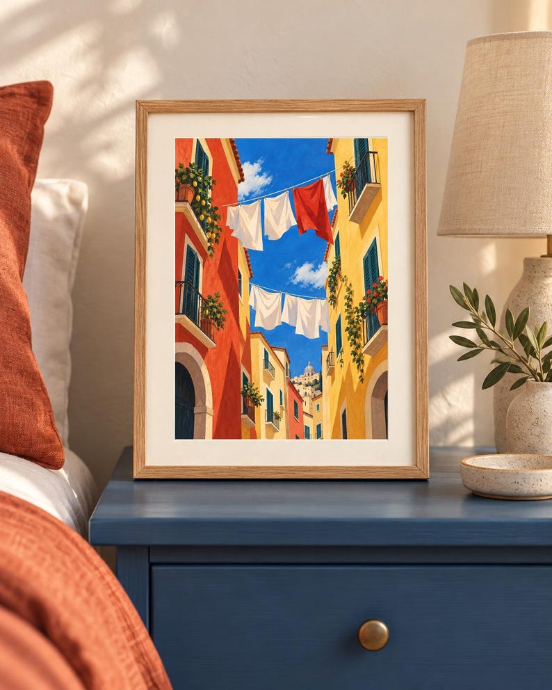Op de afbeelding: Een ingelijste kunstdruk toont een smalle straat tussen hoge oranje en gele gebouwen onder een helderblauwe lucht. Tussen de gebouwen hangt wasgoed aan lijnen, waaronder witte en rode stoffen. Potplanten sieren de balkons van de gebouwen.