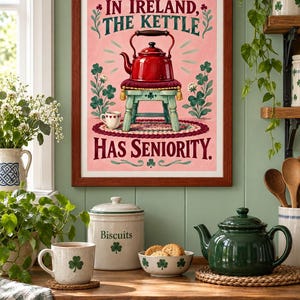 May include: A framed wall print with a pink background featuring a red kettle sitting on a small stool. The text reads, IN IRELAND, THE KETTLE HAS SENIORITY. The illustration includes green shamrock plants, a small cup, and a braided rug design.