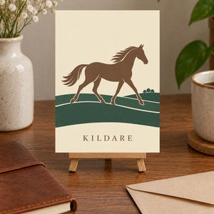 May include: A rectangular print on a small wooden easel featuring a brown horse silhouette galloping across dark green rolling hills against a cream background. The word KILDARE is printed in dark brown capital letters at the bottom of the print.
