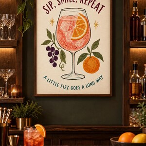 Op de afbeelding: Een ingelijste kunstprint met een centrale illustratie van een cocktailglas gevuld met een roze drankje, ijsblokjes en een schijfje sinaasappel. De tekst luidt bovenaan SIP, SMILE, REPEAT en onderaan A LITTLE FIZZ GOES A LONG WAY.