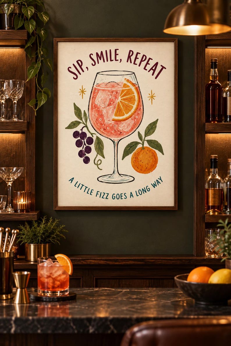 Op de afbeelding: Een ingelijste kunstprint met een centrale illustratie van een cocktailglas gevuld met een roze drankje, ijsblokjes en een schijfje sinaasappel. De tekst luidt bovenaan SIP, SMILE, REPEAT en onderaan A LITTLE FIZZ GOES A LONG WAY.