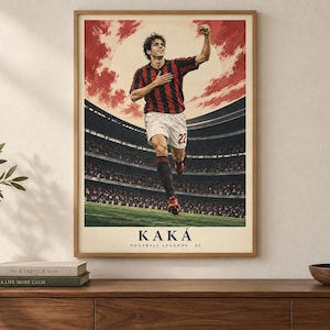 May include: A framed poster featuring a soccer player in a red and black striped jersey and white shorts, celebrating on a field with a stadium background. The text KAK&Aacute; and FOOTBALL LEGENDS 02 is printed at the bottom in a dark blue font.