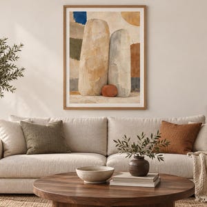 May include: A framed wall art print featuring an abstract composition of two large, vertical, stone-like shapes in beige and gray, accented by a small terracotta sphere at their base, set against a background of muted blue, green, and brown patches.