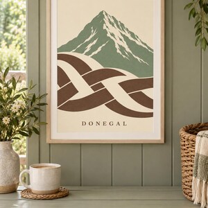 K&ouml;nnte beinhalten: Ein gerahmter Kunstdruck mit einem stilisierten gr&uuml;nen Berggipfel mit wei&szlig;en Schneeakzenten &uuml;ber einem abstrakten braun-cremefarbenen geflochtenen Knotenmuster. Das Wort DONEGAL ist in einer einfachen Serifenschrift auf dem cremefarbenen Hintergrund gedruckt.