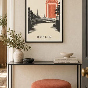K&ouml;nnte beinhalten: Ein gerahmter Wandkunstdruck mit einer stilisierten Illustration von Dublin, Irland. Die Grafik zeigt eine schwarze Silhouette einer Br&uuml;cke und einer Skyline vor cremefarbenem Hintergrund, mit einer markanten orangefarbenen T&uuml;r und dem Text DUBLIN.