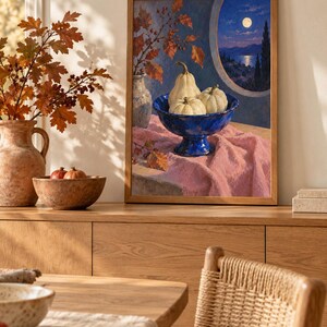 May include: A framed wall art print depicting a still life scene with three pale pumpkins in a deep blue pedestal bowl, set on a pink cloth. The background shows a circular window view of a moonlit sea, dark hills, and autumn foliage in a vase.