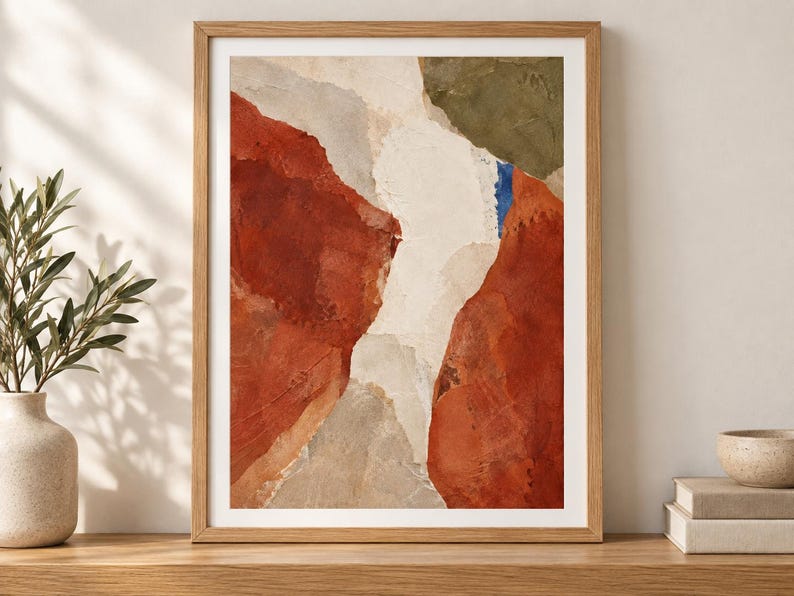 May include: A framed abstract wall art print featuring layered shapes in terracotta, cream, olive green, and a small accent of blue. The composition displays textured, organic forms with visible brushstroke details enclosed in a light wood frame.