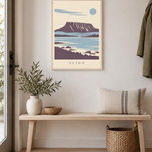 May include: A rectangular wall art print in a light-colored wooden frame featuring a stylized landscape illustration of a mountain, water, and shoreline. The print has a cream background, blue sky with a circular sun, and the text SLIGO at the bottom.