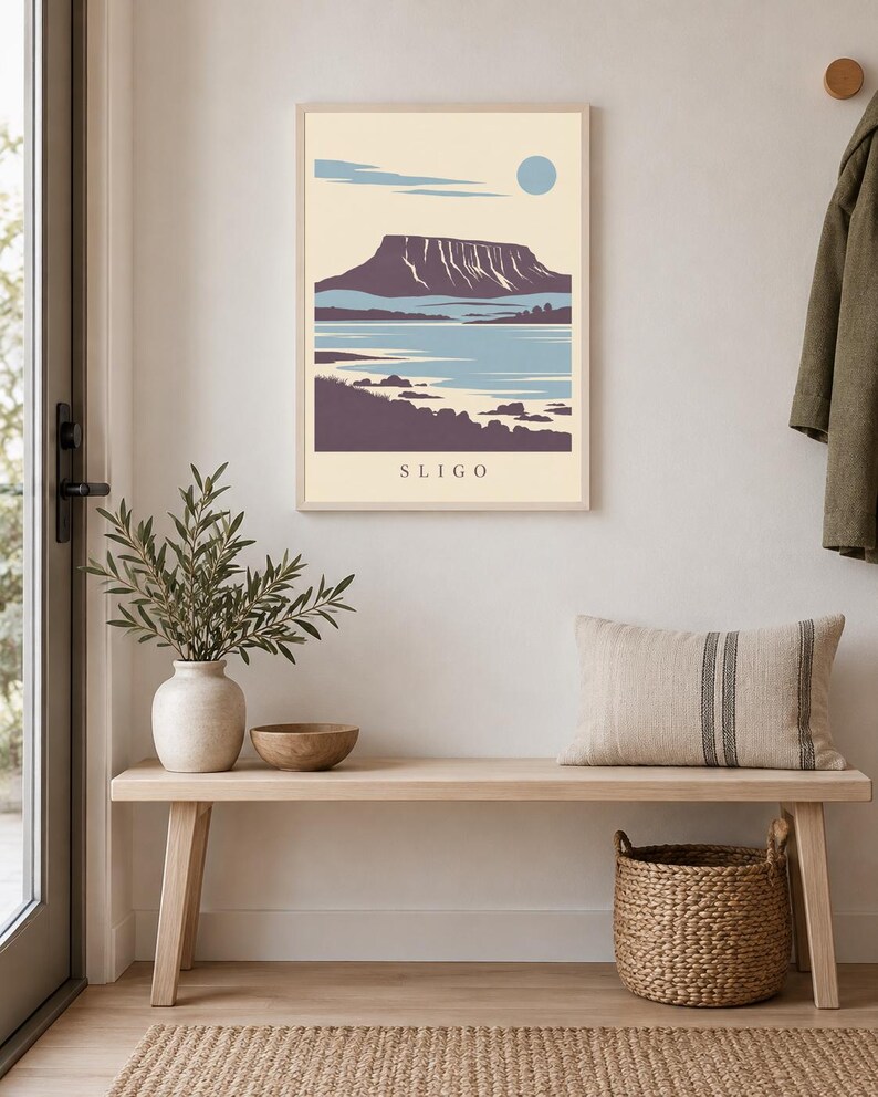May include: A rectangular wall art print in a light-colored wooden frame featuring a stylized landscape illustration of a mountain, water, and shoreline. The print has a cream background, blue sky with a circular sun, and the text SLIGO at the bottom.